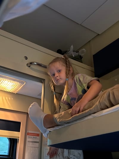 The image shows a young girl with blonde pigtails and blue eyes looking up at the camera. She is wearing a tie-dye t-shirt and a white sock. The girl appears to be in a train compartment, indicated by the bunks and the general interior design. One bunk is visible above her, with someone's legs and feet, wearing a white sock, resting on the edge. The lighting suggests it might be daytime, possibly evening, given the warm glow from a light fixture. The scene is likely set within Russia, as a Cyrillic text label is visible on the wall, which reads "Росплансированный" and "Саратов - Москва" (Rosplanrovannyy and Saratov - Moscow), indicating a train route. The girl's expression is curious and perhaps a little playful. The overall impression is one of a cozy and intimate moment within a travel setting.