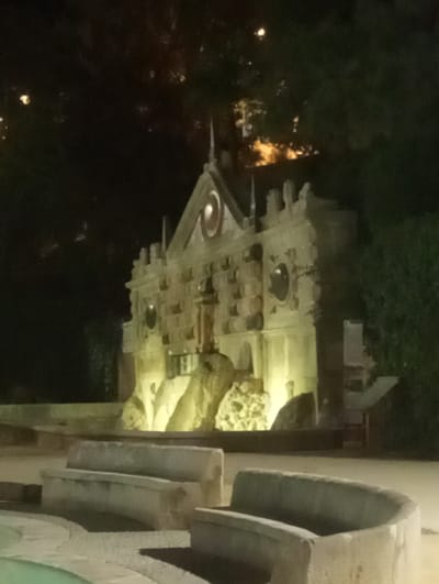 The image shows a nighttime view of a decorative fountain facade with stone benches in the foreground. The fountain structure is illuminated with warm yellow light, highlighting its intricate carvings and architectural details, including what appears to be an eye-shaped motif at the top. Natural elements, such as trees and foliage, form a dark backdrop against the illuminated structure. The stone benches are empty and appear weathered. This fountain is identified as the Fuente del Rey in Priego de Córdoba, Spain.