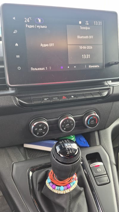 This media file captures a close-up view of a car's interior, focusing on the center console and dashboard. The primary objects visible are a car's infotainment touchscreen, climate control knobs, a gear shifter, and the electronic parking brake button. The infotainment screen displays information related to the phone and audio system, showing "Телефон" (Phone), "Bluetooth OFF," and the date "10-06-2026" with the time "13:31." It also shows the temperature "24°C" and "Радио / музыка" (Radio / Music), along with "Аудио OFF" (Audio OFF).
The climate control area features three rotary knobs, one for A/C, another for fan speed or temperature settings (indicated by numbers 0-4 and symbols), and a third with symbols for defrosting and airflow. Below these knobs are various buttons, including an "ECO" button and hazard lights.
The gear shifter is a prominent feature, with a manual transmission pattern clearly visible on its top. It is adorned with colorful bracelets made of beads and rubber bands. In the background, the electronic parking brake button and what appears to be a storage compartment containing a blue object and a green item (possibly a lighter) are visible.
The scene is indoors, within a car, suggesting a daily commute or a parked vehicle. The lighting appears to be natural daylight, indicating it could be daytime. There are no people visible in the image. The text present is in Russian and appears to be standard car interface text. The location context is Preiļi, Latvia, which provides a geographical setting for the image.