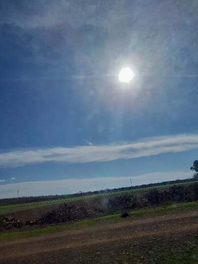 The scene depicts a rural landscape in Oued Jdida, Morocco, under a partially cloudy blue sky. The sun, prominent in the upper right quadrant, casts strong light and distinct lens flares across the frame. The foreground comprises a section of brown earth or a dirt path, sloping gently upwards from the lower right, containing scattered small stones and patches of green vegetation. In the midground, a substantial pile of dark, irregularly shaped rocks occupies the left half, situated between the foreground and agricultural fields. These fields display both tilled brown soil and areas of vibrant green crops. The background extends with additional green cultivated areas to the horizon, where a distant line of darker foliage, possibly trees or bushes, is visible. A slender utility pole stands erect in the distant left-center, and the crown of a tree is partially visible on the far right edge of the frame. No human figures or animals are present.
