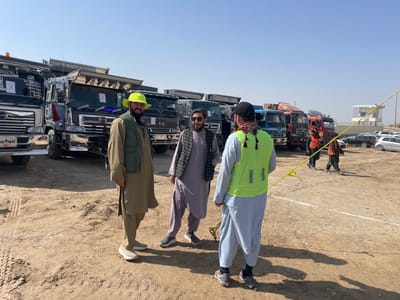 The image depicts an outdoor scene in Pir Dust, Afghanistan, under a clear blue sky. In the foreground, three adult males are engaged in conversation on dry, unpaved ground. The man on the left wears a light brown tunic, a matching vest, a bright yellow bucket hat, and light-colored sneakers. The central man wears a light purple tunic with a dark patterned vest and dark sneakers. The man on the right, viewed from the back, wears a black baseball cap, a light blue tunic, a yellow-green high-visibility vest, a scarf with black tassels, and dark sneakers.
In the mid-ground, behind the three foreground individuals, a line of multiple large commercial dump trucks, predominantly Hino models with chrome accents and personalized details (e.g., "Samir Popal" on one windshield), are parked in rows. A yellow rope or cord extends across the ground from the mid-right towards the left. Further back and to the right, two additional men, both wearing orange high-visibility vests, walk away from the camera. The background reveals a simple, light-colored building, possibly a guard tower or office, and several parked passenger cars.