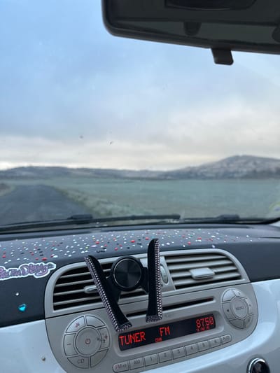 The image provides a view from the interior of a vehicle, looking out onto a rural or semi-rural landscape within Frontenas, France. A paved road extends into the distance, flanked by fields that appear covered in a light frost or winter vegetation. In the background, undulating hills are visible under an overcast, grey sky.

Inside the vehicle, the black dashboard is customized with numerous small, multi-colored dots (including red, white, pink, blue, yellow, orange, green, and black) and two stickers on the left: a purple and white text sticker, and a small blue glitter heart sticker. A black phone holder, embellished with small rhinestones, is clipped to one of the central air vents. Below it, a white car radio console is visible, displaying "TUNER FM," "87.50" (frequency), and "CD-IN." The base of the windshield wipers and the lower edge of the rearview mirror are also present. The dashboard design and radio unit are consistent with a Fiat 500 vehicle. No individuals are visible in the scene.