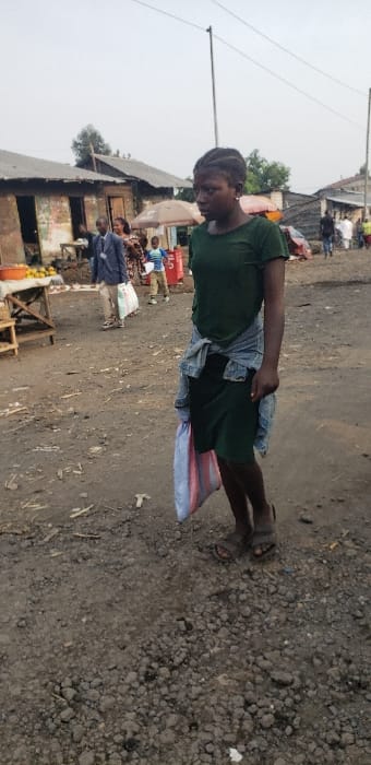 An outdoor scene in Buzove, Democratic Republic of the Congo, depicts a busy, unpaved street or market area under an overcast sky. In the foreground, a young female, approximately 18-25 years old, walks towards the viewer's right. She wears a dark green short-sleeved shirt, a matching dark green knee-length skirt, and a light blue denim jacket tied around her waist. She is wearing dark sandals and has dark hair styled in braids. In her left hand, she carries a large, vertical striped bag in shades of blue, white, and pink.

The ground is composed of dark, loose dirt and gravel, with various small pieces of debris scattered across it. In the midground and background, multiple individuals are visible. To the left of the primary subject, a man in a light blue suit jacket and dark trousers walks with a white bag. Other figures, including a young child in a blue shirt, are present further back. Structures in the background consist of single-story buildings with corrugated metal roofs and weathered walls, some appearing to be market stalls. A wooden stall displaying yellow-orange produce is visible to the immediate left of the frame. Several utility poles with overhead wires extend into the sky. Other market elements, such as brown vendor umbrellas and a red container, are also present. The scene indicates a populated commercial or residential area.