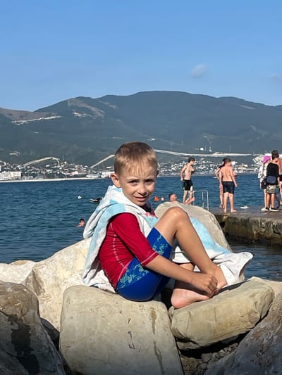 A young boy is resting on rocks near the sea. He is wearing red swimwear and is wrapped in a light blue and white towel. His legs and feet are visible, with one foot tucked under him. In the background, other people are in the water and on a concrete pier. The sea is a deep blue, and a town with buildings is nestled against a green, mountainous landscape. The sky is clear and blue, suggesting it is a sunny day, likely in the afternoon. The overall scene conveys a relaxed atmosphere of a day at the beach or seaside resort in Ol'ginskii, Russia.
