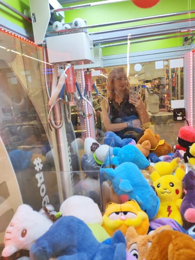 In Vasil'ki, Russia, a woman with reddish-blonde hair is taking a selfie in the mirrored glass of a claw machine arcade game. The game is filled with an assortment of plush toys, including a large orange Garfield, a yellow Pikachu, a blue whale, and a pink Hello Kitty. The woman is wearing a black lace top and several silver bracelets. The claw machine itself is white with red accents and appears to be operational. The background reflected in the glass shows a store aisle with shelves of products, suggesting the arcade is part of a larger retail establishment.