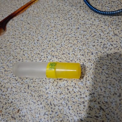 The image shows a close-up, eye-level, horizontal shot of a translucent cylindrical tube with a yellow cap, lying on a speckled, grey and tan surface. A small, green, leaf-like pattern is visible on the yellow cap. To the left of the tube, a portion of a dark brown and orange comb is visible. In the upper right corner, a dark blue, ribbed cable is partially visible. The surface appears to be a countertop or table with a textured, speckled pattern. No people or actions are depicted. The lighting suggests it is indoors, possibly in a kitchen or bathroom setting, but its specific location within Reudeuep, Indonesia, cannot be determined from the visual information.