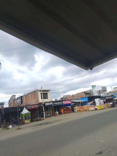 The image depicts a street scene in Gampongpaya, Indonesia, on an overcast day. In the foreground, a dirt road with scattered debris runs diagonally across the frame. Along the right side of the road, a row of shops and stalls is visible. These establishments feature various signs, including "AURISTYLE," "MAYA modo," and "WM BOUTIQUE." One stall has a green and white tent and displays advertising for "ES TELER KANTARARA." Further down the row, colorful banners advertise "ES TELOR CREAM DURIAN." Several motorcycles are parked in front of the shops, and a few people are standing or walking. In the background, buildings with brick facades and corrugated metal roofs are present. Overhead, a large, dark-colored structure, possibly a canopy or overhang, partially obscures the sky, which is filled with dramatic, grey clouds.