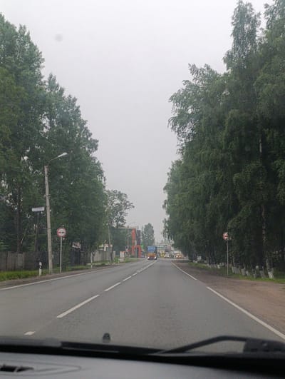 The media file shows a view from inside a vehicle driving on a road lined with tall trees on both sides. The sky is overcast, suggesting a cloudy or foggy day. The road is paved and has a white dashed line marking the lanes. In the distance, there are some buildings and vehicles, including a large yellow truck.
Notable details include a street lamp and a road sign on the left side of the road. The sign appears to be a circular red sign with a white horizontal bar, possibly indicating a restriction. Another sign with Cyrillic text is attached to the lamppost. On the right side of the road, a similar red and white circular sign is visible. The presence of trees and buildings suggests a semi-urban or rural setting. The overall mood is subdued due to the overcast weather.
The visible text on the sign attached to the lamppost appears to read "777-3-506" and below it some smaller Cyrillic text, likely a street name or address.