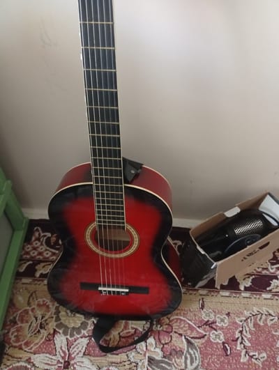 An acoustic guitar with a red and black finish stands vertically on a patterned rug. The guitar is in the foreground and is the primary subject. The background consists of a plain, light-colored wall. To the right of the guitar, a cardboard box contains what appears to be a black microphone and its accessories. A green object, possibly a piece of furniture or shelving, is partially visible on the left side of the image. The scene is indoors, with the lighting suggesting it is daytime. There are no people visible in the image. The image is a still photograph with no observable action. The location in the city of Seýdi, Turkmenistan is not discernible from the visual information provided in the image.