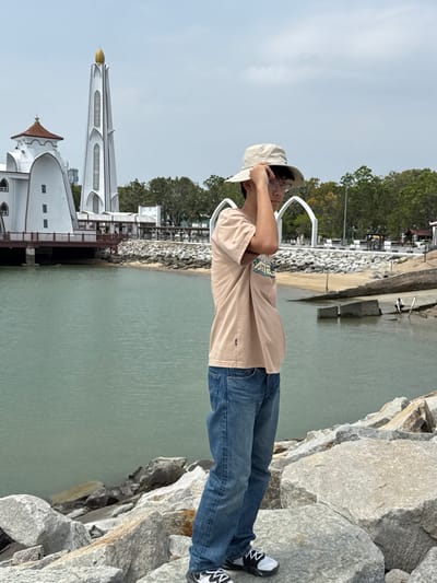 A person in a beige bucket hat, tan shirt, and blue jeans stands on large rocks near the water. Behind them, a beach and bridge-like structure are visible. To the left, an architectural building with a pointed tower stands on the water, connected by a boardwalk. The sky is overcast.