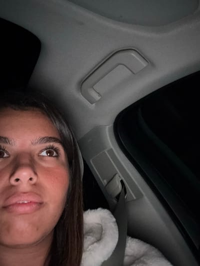The image depicts a partial, upward-angled view inside a vehicle at night, reportedly located in West Windsor, United States. No specific geographical identifiers for West Windsor are visually present in the image.
A female individual is visible on the left side of the frame, showing her face from the upper lip to the forehead. She has dark brown hair, brown eyes with prominent eyelashes, and a warm-toned complexion with a reddened area on the right cheek. Her gaze is directed upwards and slightly to the right. She is wearing a light-colored, textured garment, possibly faux fur, and is secured by a dark grey seatbelt that crosses her lower right torso.
The vehicle's interior features a light grey headliner, a light grey overhead grab handle mounted above the A-pillar, and light grey A-pillar trim. The exterior environment visible through the window is uniformly dark, consistent with nighttime conditions. The interior is illuminated, causing reflections in the individual's eyes.