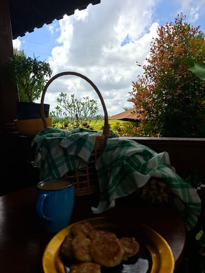 The image shows a view from what appears to be a balcony or porch in Lodtunduh, Indonesia. The camera is tilted. In the foreground, on a dark surface, is a gold-colored plate piled with what appear to be fried patties. Next to the plate is a blue mug and a woven basket draped with a green and white checkered cloth. The basket contains a pineapple. Through the basket's handle and the gap between the railing and roof, the outdoor scene includes a partly cloudy blue sky and trees with red and green foliage. Parts of a roof are visible at the bottom left. Overhead electrical lines are also visible.