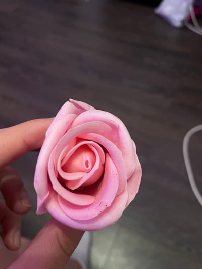 A close-up shot focuses on a pink, artificial rose held between a person's thumb and index finger. The rose exhibits tightly curled petals, creating a spiral pattern towards its center. The background is blurred, showing a dark wooden surface and some out-of-focus white and purple objects. The lighting is soft, highlighting the delicate texture of the rose and the skin of the finger. No specific location in Romanovka, Russia is identifiable from the image.