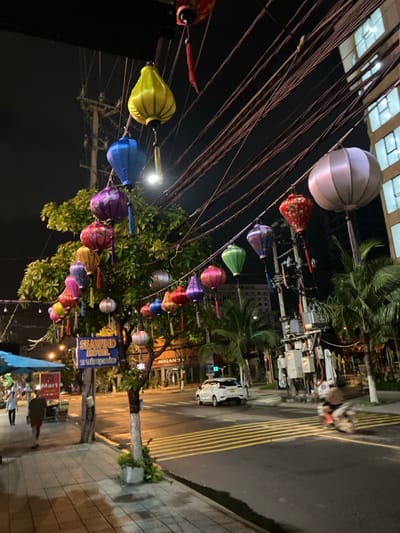 The image depicts a nighttime street scene in Ngũ Hành Sơn Ward, Vietnam. The foreground features a concrete sidewalk with two pedestrians; one individual in a dark shirt and red shorts walks away from the camera, while another in a lighter shirt is partially visible further down. To the left, a blue awning covers an outdoor stall, and a red "Mart" sign is visible. A tree central to the sidewalk is heavily decorated with numerous multicolored silk lanterns of various shapes, including yellow, blue, purple, red, green, and silver. A blue sign for "SEAWIND HOTEL, 10 TRẦN BẠCH ĐẰNG" is attached to the tree.
The midground shows a paved two-lane road with distinct yellow horizontal striped markings. A white passenger vehicle is parked on the right side of the road, and a motion-blurred scooter with a rider is traversing the road from right to left. Overhead, a dense network of black electrical wires crisscrosses the dark night sky, with additional lanterns strung between poles and across the street. A street light is also visible. In the background, a multi-story building with illuminated windows stands on the right, and palm trees are interspersed along the street and near the buildings. A "HIGHLANDS COFFEE" sign is visible in the distance.