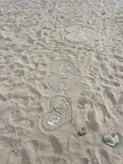 In the city of Karolino-Buhaz, Ukraine, on a sandy beach, the word "FOREVER" is written in seashells and small stones arranged in a heart shape. The word "LOVE" is also written in seashells, positioned above "FOREVER" and within another heart shape. The scene is illuminated by natural daylight, casting subtle shadows on the textured sand. The surrounding environment is a natural beach with scattered shells and pebbles. There are no people visible in the image.