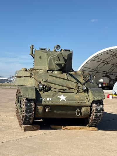 An olive drab military tank is positioned on concrete. The vehicle, marked with a white star and the inscription "2Δ 87," exhibits signs of wear and rust. It has tracks and a prominent gun turret. A large radar or satellite dish structure looms in the background under a clear blue sky. Wood blocks are positioned under the tank's tracks.