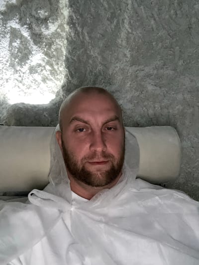 A man with a shaved head and a beard lies on a light-colored pillow in a salt cave. He is wearing a white disposable suit. The cave walls are textured and appear to be made of salt or a similar mineral. The lighting is dim, with a bright light source visible in the upper left corner. The man is looking directly at the camera.