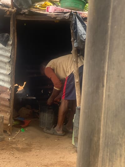 In Idiroko, Nigeria, a rustic, open-front shelter constructed from corrugated metal sheets and wooden supports features a dark interior. An adult male, with dark skin, is visible within the structure, bent forward at the waist. He wears a beige t-shirt over reddish-brown long sleeves, navy blue shorts with a red vertical stripe, and grey slip-on sandals. His hands are positioned over one of two dark, cylindrical buckets on the dirt ground inside the shelter. Further back in the dark interior, a black pot rests on a stand over a visible heat source, emitting light and light smoke. The shelter's makeshift roof area supports a green plastic bucket, a pink plastic dish rack, and a yellow fabric item. The immediate ground outside the shelter is bare, dry earth. To the far right, a large, light grey corrugated sheet or wall partially obscures the scene, with a wooden support pole positioned in front of it.