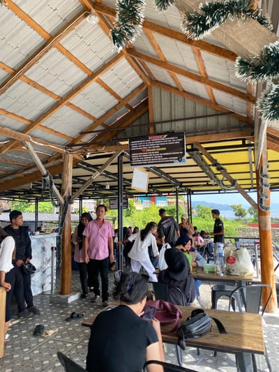 The image depicts an outdoor seating area with a thatched roof supported by wooden beams. Several people are present, some standing and others seated at tables. The setting appears to be a restaurant or cafe in Tuk Tuk, Indonesia, with a body of water and lush greenery visible in the background. One man in a pink shirt and black pants is looking towards the camera, while other individuals are engaged in conversations or browsing their phones. The floor is tiled, and several pairs of sandals are scattered around, suggesting a relaxed atmosphere. Decorative tinsel is hanging from the wooden beams, possibly for a festive occasion.