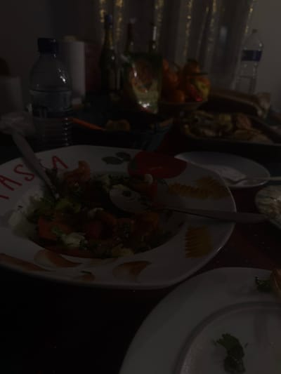 A dimly lit dining table features an arrangement of plates and food items. A salad with greens and tomatoes occupies a decorative plate with lettering. Beside it, a clean white plate contains a small green garnish. Further back, a bowl of oranges sits near bottles and glasses. Utensils are scattered amongst the dishes, suggesting a meal in progress. The composition is set against a dark backdrop.