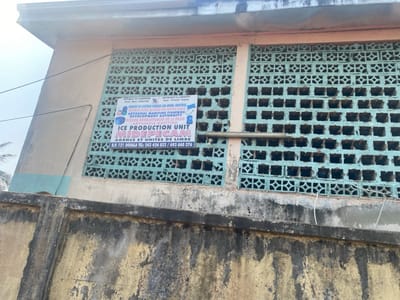 An outdoor, eye-level shot captures a portion of a building, likely a utilitarian or industrial structure, possibly in Victoria (Limbe), Cameroon. The building features a light-colored (beige or cream) facade and a section of two large rectangular window openings filled with decorative, light blue-green concrete breeze blocks. These blocks serve as ventilators or sunscreens. A metal pipe runs horizontally across the upper part of these blocks.
A white rectangular sign with blue text and logos is prominently displayed on the breeze block section of the building. The sign indicates the presence of a "REPUBLIC OF CAMEROON" entity, specifically the "MINISTRY OF LIVESTOCK FISHERIES AND ANIMAL INDUSTRIES" (and its French equivalent). It identifies the facility as an "ARTISANAL MARITIME FISHERIES DEVELOPMENT AUTHORITY / MISSION DE DEVELOPPEMENT DE LA PECHE ARTISANALE MARITIME," and more specifically, an "ICE PRODUCTION UNIT" for "MIDEPECAM." The sign also provides the location as "AGENCE ET UNITES DE LIMBE" (Agency and Units of Limbe), along with a post office box number for Douala and telephone numbers: 243 424 033 / 693 660 376.
In the foreground, a stained, weathered concrete wall rises, showing signs of dirt and possibly mildew, suggesting an older structure in a humid environment. The building itself appears somewhat weathered, with marks and discoloration. The sky visible above the building is bright but appears hazy or overcast, indicating a daytime shot without direct sunlight. There are no people visible, and no active events are taking place. The overall impression is a functional, albeit aged, facility.