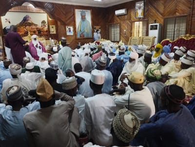 The scene is set in a large room with wood-paneled walls and a high ceiling. There are approximately 40 people seated in rows on the floor. Most of the people are men, wearing traditional white or light-colored clothing and caps. Some women are seated in the back, and a few children are present. The men are engaged in a conversation or a meeting. In the back of the room, there are several gold-framed portraits on the wall, including a large portrait of a man in a white robe. Several air conditioning units are on the walls, and a large, ornate chandelier hangs from the ceiling. The image was taken in the Emir’s Palace in Kaltungo, Gombe State, Nigeria. The coordinates given place the image in the palace's courtyard area, which is a place used for community gatherings, meetings, and ceremonies.