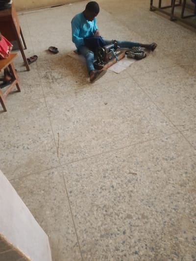 In Rumuekini, Nigeria, a single male individual is depicted operating a manual sewing machine while seated on the tiled floor. The man, wearing a light blue long-sleeved shirt and blue jeans, is positioned centrally, facing downwards towards the sewing apparatus. He is seated on a light-colored mat, with his legs extended forward, his feet clad in dark flip-flops. A dark-colored sewing machine, mounted on a wooden base, rests on a similar light-colored mat directly in front of him. His hands are engaged with a dark blue fabric piece being sewn by the machine. To the man's right, a patterned, dark-colored pouch or bag is visible on the floor. Two additional pairs of dark flip-flops are situated on the floor to the left, near a wooden stool with a reddish item on its seat. The floor consists of light grey terrazzo tiles with visible grout lines and minor dark debris. The background features a light-colored wall and the partial view of metal-framed furniture.