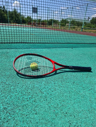 A tennis racket and ball lie on a green tennis court. The racket is red and black with white strings, and the ball is yellow. A black tennis net is visible in the background, with trees and a building behind it. The sky is blue with some clouds. The image appears to be taken on a sunny day. There are no people visible in the image. The location is identified as Gorkovskiy, Russia.