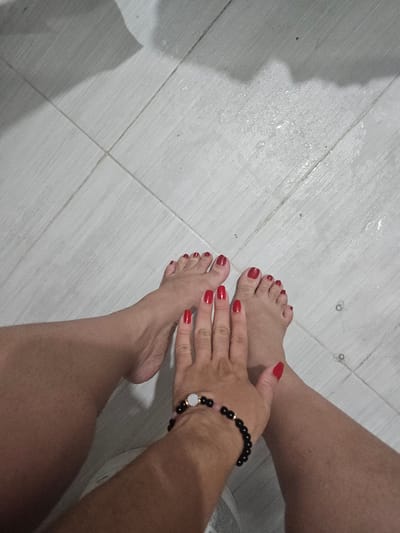 A top-down view shows a woman's feet and hands on a light gray tiled floor, possibly in Perímetro Urbano Cúcuta, Colombia, though no location information is visible in the image. Her toenails and fingernails are painted bright red. She is wearing a black beaded bracelet on her wrist with a white and gold decorative element. The floor appears slightly wet in certain areas.