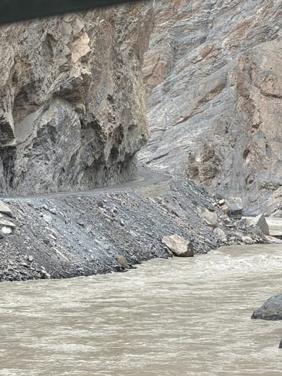 The image depicts a winding road carved into the side of a steep, rocky mountain overlooking a fast-flowing, muddy river. The road appears to be made of gravel or dirt and hugs the cliff face, with loose rocks and scree lining its edge. The mountain itself is composed of layered, grey rock formations, indicating a significant geological history. The river below is wide and turbulent, with visible ripples and white water suggesting a strong current. The environment appears rugged and remote, characteristic of a mountainous region. The image does not contain any people or specific landmarks that would allow for precise location identification within Stongde, India, beyond its general mountainous and riverine landscape.