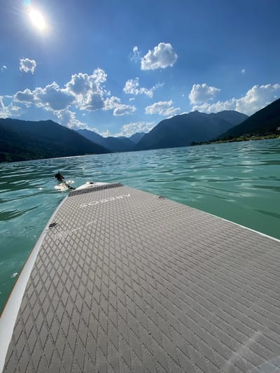 The image captures a first-person perspective from a paddleboard on a serene lake, likely in Lärchenwiese, Austria, given the mountainous backdrop and clear blue sky with scattered white clouds. The sun, a bright, almost oval shape, shines intensely in the upper left corner, casting light rays that pierce through the clouds. The water is a vibrant turquoise, with gentle ripples reflecting the sunlight. In the foreground, the textured grey surface of the paddleboard is visible, featuring a diamond-shaped pattern. A black paddle is partially submerged in the water to the left, connected by a rope to the board. The background is dominated by dark, imposing mountains covered in lush green trees, their slopes descending towards the lake. On the far side of the lake, a thin strip of land with some buildings and scattered trees can be observed. The overall atmosphere is tranquil and idyllic, suggestive of a peaceful summer day spent on the water.