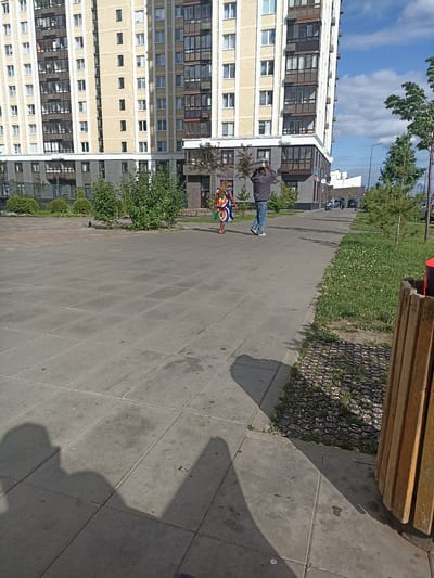 In Ostafievo, Russia, a contemporary apartment building with a cream-colored facade and numerous windows dominates the background of this outdoor scene. In the mid-ground, two individuals are walking on a paved pathway in front of the building. One person, dressed in jeans and a gray jacket, is facing away from the camera with their hands raised to their head. The other individual, wearing a colorful dress, is walking to the left of the first person. To the right of the pathway, a patch of green grass with small white flowers is visible, adjacent to a wooden trash receptacle. The foreground consists of a large paved area with square tiles, casting shadows from unseen objects. The sky above is partly cloudy.