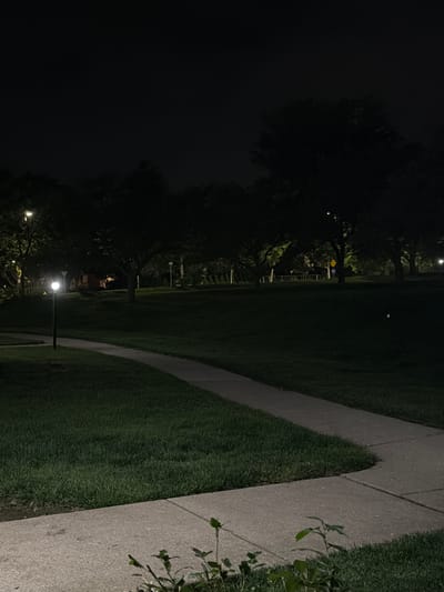 In the city of Vernon Hills, United States of America, a park path is illuminated by a single lamppost on a dark night. The path, made of concrete, curves through a grassy area. Trees are silhouetted against the dark sky, some with faint light emanating from behind them. In the distance, a yellow sign is visible. The overall scene is one of a quiet, dimly lit park at night.