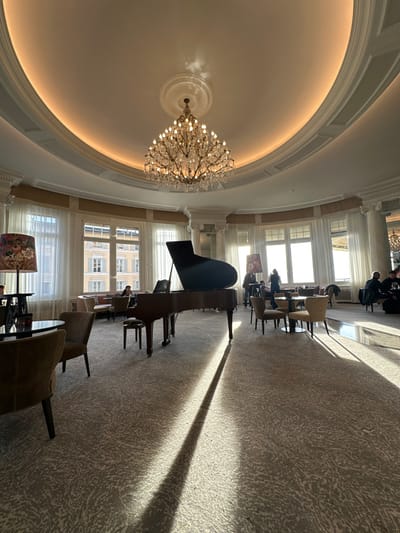 This image depicts the opulent lobby lounge or bar area of the Beau-Rivage Palace, located in Ouchy, Lausanne, Switzerland. The large, elegant room features a high, curved ceiling with decorative coving and a prominent, illuminated crystal chandelier suspended centrally. Multiple tall windows, adorned with sheer white curtains, line the exterior wall, providing a view of traditional multi-story urban buildings across a street.

A dark brown grand piano with its lid open is centrally positioned on a patterned light gray carpet, accompanied by a matching piano stool. Strong natural sunlight streams in from the windows on the right, casting long, distinct shadows across the carpet from the piano and other furniture.

Various seating arrangements are dispersed throughout the room. To the left, two light-upholstered armchairs flank a small round table. Further back on the left, multiple plush armchairs and a sofa are arranged around low tables. On the right side, additional light-upholstered armchairs are grouped around small round tables. Tall decorative lamps with patterned fabric shades are visible on tables in the foreground left and mid-ground right.

Several individuals are present: one person is seated on a sofa in the far background on the left. Towards the right side of the room, a group of approximately three to four individuals are either standing or seated around tables, appearing to engage in casual conversation or waiting. Two additional individuals are seated at a table on the far right. Ornate classical columns separate sections of the windows.