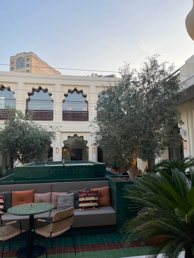 This image depicts an outdoor courtyard located within The Chedi Al Bait, Sharjah, United Arab Emirates.
The scene presents an open-air area surrounded by a cream-colored building featuring traditional Emirati architectural elements. The upper level of this building displays a series of identical scalloped arch windows, each fitted with dark wooden lattice railings. The lower level includes similar arched recesses, flanked by rectangular modern wall sconces. In the distant background, a taller, lighter tan building with a prominent large arched window near its peak is visible against a clear, light blue sky. Overhead, thin electrical wires traverse the upper section of the main building.
The courtyard's foreground and midground are structured around a multi-tiered, glossy dark green tiled water feature or planter. Integrated into this structure is an L-shaped built-in sofa with grey cushions and an assortment of throw pillows in shades of orange, terracotta, cream, and patterned fabrics. In the immediate foreground, a small, round table with a green speckled surface rests on a dark pedestal base, accompanied by two chairs featuring light-colored woven seats and backrests on dark metal frames.
Several olive trees with dense, silvery-green foliage are positioned throughout the courtyard, some planted in matching square dark green tiled planters. Smaller, dark green, palm-like shrubs are also visible in the foreground. The ground is paved with a mosaic of small square tiles in varying shades of dark green, teal, and reddish-brown.
In the deeper midground, behind a prominent olive tree, a few indistinct figures are seated at an additional outdoor seating arrangement, appearing to be patrons. The overall lighting suggests late afternoon or early evening conditions, characterized by soft, diffused illumination.