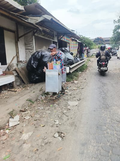 The image captures a bustling roadside scene, likely in an urban or semi-urban setting, characterized by informal commerce and daily activity under an overcast sky.

In the foreground to the left, a male vendor stands behind a small, metallic cart, which appears to be used for selling drinks or snacks. He is wearing a dark baseball cap, a plaid long-sleeved shirt (featuring red, blue, and white squares), and dark trousers with flip-flops. His right arm is bent, and his hand is near his face, suggesting he might be taking a sip of a drink, eating, or making an adjustment to his cart. On top of the cart are several plastic containers holding various liquids, some with straws and small utensils or skewers sticking out.

Behind the vendor and his cart, against the backdrop of makeshift structures, a large pile of black plastic trash bags is visible, overflowing and stacked against a low metal road barrier. These structures, likely stalls or small shops, are made from a combination of wood, corrugated metal sheets for roofing, and possibly tarpaulin for walls, indicating an informal retail area. Further down the row of stalls, one structure displays a colorful arrangement, possibly products or decorations.

To the right of the vendor, an asphalt road stretches into the distance, showing signs of wear with cracks and patches. A black scooter or motorcycle, ridden by a person wearing a dark helmet and a light-colored backpack, is seen traveling away from the viewer on this road. The motorcycle's license plate "N 3427 EUG" is partially visible, with "08-25" (likely an expiry date) below it. In the far background along the road, more vehicles and structures are discernible, and a large concrete bridge or overpass can be seen spanning across the scene.

The ground area between the stalls and the road is unpaved, consisting of dirt and gravel, and is littered with various debris, small pieces of trash, and sparse patches of green vegetation. The sky above is largely cloudy, suggesting an overcast day, but the natural light indicates it is daytime. The overall atmosphere conveys an active, lived-in environment where street vending is a common sight amidst somewhat untidy surroundings.
