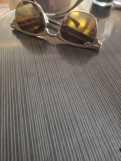 A pair of brown-tinted sunglasses, with a silver metal frame, rest on a gray, ribbed surface. The sunglasses have a slight curve to their frames and are positioned with the right lens facing the viewer. The surface appears to be a table or countertop. The location is a private residence in Tunis, Tunisia, based on the provided coordinates. There are no people or other objects visible in the image.