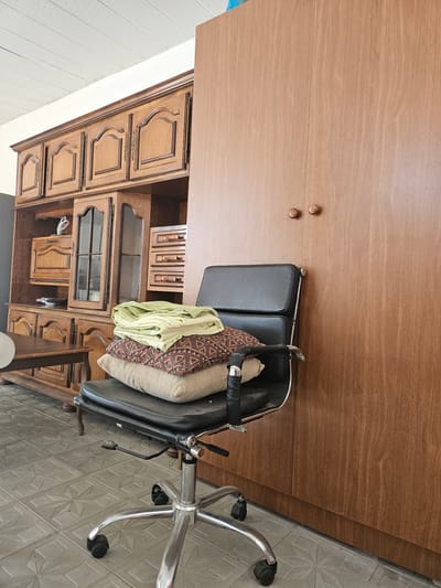 The image depicts an indoor scene, likely in a room in Saray, Azerbaijan. A dark leather office chair with chrome accents is positioned in the foreground. The chair is occupied by a stack of three folded textiles. The lowest textile is a beige cushion. The middle textile is a patterned cushion with geometric designs in brown, red, and black. The top textile is a folded light green sheet or blanket.
Behind the chair is a large, dark wooden hutch or cabinet unit. This unit features multiple doors and drawers, with decorative carved details on some of the upper doors. The hutch has glass-fronted cabinets on the left side, revealing some items within. To the right of the hutch, a tall, plain wooden cabinet door is visible, with two small wooden knobs near the top.
The flooring is made of grey square tiles with an embossed geometric pattern. In the background, part of a white ceiling with a textured surface is visible. A dark wooden table is partially visible to the left of the chair. The overall impression is of a storage or office area that is somewhat cluttered.
