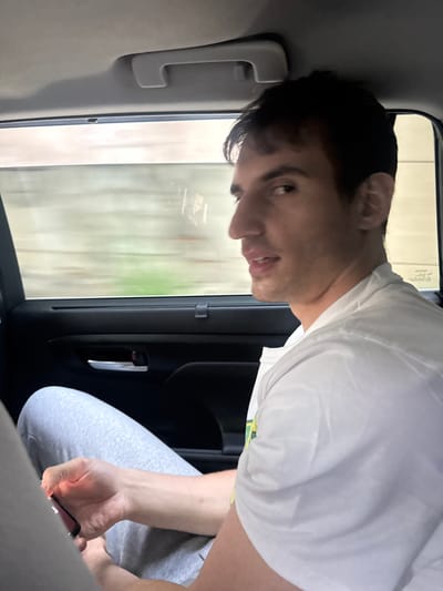 A young adult male is seated in the rear passenger compartment of a vehicle, positioned in the city of Fort Lee, United States. He has short dark hair and is wearing a white t-shirt and light gray sweatpants. His left hand is holding a dark-colored smartphone. He is turned towards the viewer, with his mouth slightly ajar, looking directly at the camera. Through the car window, a blurred, light-colored background consistent with a tunnel wall, exhibiting motion streaks and faint greenish hues, indicates the vehicle is in motion. The car interior shows a gray overhead grab handle and a black door panel with a silver door handle.