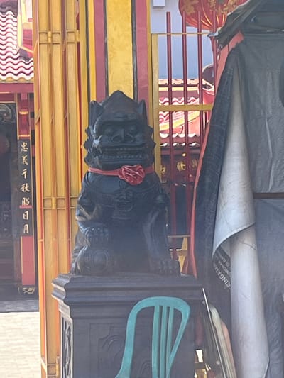 In Desa Tambakreja, Indonesia, a black stone statue of a lion-like creature, possibly a guardian or temple lion, stands prominently on a dark pedestal. The statue is adorned with a red collar that has a red floral embellishment. Behind the statue, a vibrant yellow and red structure with vertical bars is visible, suggesting it might be part of a temple or traditional building. A light-colored rolled-up tarp or fabric is draped to the right of the statue, obscuring a portion of the background. To the left of the statue, a black column features vertical script in white, possibly Chinese or other Asian characters. A bright turquoise chair is placed in front of the pedestal, partially obscuring it from view. The ground in front of the structure appears to be paved with light-colored square tiles. The overall scene suggests a cultural or religious site within the village.