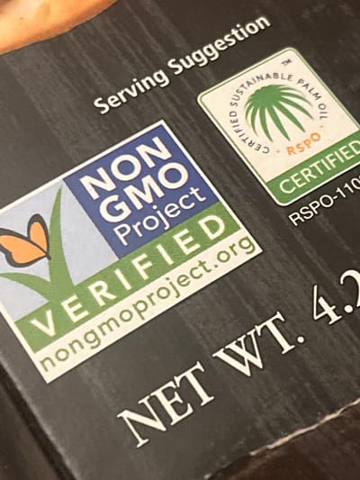 This image captures a close-up, angled view of a product package, predominantly dark in color. The focus is on several prominent certifications and product details printed on the surface.
In the center, a blue and green logo for the "NON GMO Project VERIFIED" is clearly visible, along with its website "nongmoproject.org" written vertically. Below this logo, the bottom portion of a monarch butterfly illustration is visible.
To the upper left of the Non-GMO label, a green and white logo for "SUSTAINABLE PALM OIL RSPO CERTIFIED" is present, featuring a palm tree icon and the certification number "RSPO-110".
Other visible text includes "Serving Suggestion" printed horizontally along the bottom left edge, and "NET WT. 4.2" in white capital letters on the upper right side of the package. The entire image is slightly rotated, giving an oblique perspective of the packaging details. The setting appears to be an indoor environment, typical for product photography.