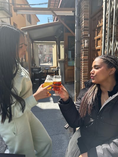 This image displays two individuals toasting with drinks on an outdoor patio in El Espinar, Spain, under clear daylight conditions. The person on the left, with long, dark hair, wears a light green tracksuit and holds a glass of amber-colored liquid in her right hand, looking towards the second individual. The person on the right, with dark braided hair, wears a black puffer jacket over a dark pleated garment and holds a glass of reddish-brown liquid in her left hand, facing the first individual with a pouting expression. Their glasses are clinking together. The background features a paved street with an outdoor seating area under an awning, a sign displaying 'Protos', and multiple traditional buildings constructed of brick and plaster. A metal security gate and a vertical downspout are visible on the right-hand building. Distant figures are faintly discernible further down the street.