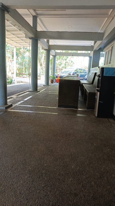 A covered outdoor seating area is depicted. The floor is made of dark, pebble-like concrete. A dark brown, rectangular table is centrally located in the foreground, with a dark blue water cooler to its right. Behind the table, there are two long, dark brown benches. A dark blue vehicle is visible in the background, parked under the covered area. An orange traffic cone is placed in front of the vehicle. The surrounding environment includes trees and foliage. The scene appears to be in Sumedang, Indonesia.
