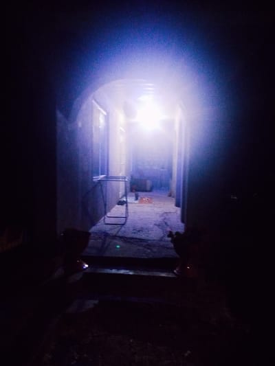 The image shows a dimly lit, narrow hallway or passage, possibly an outdoor corridor, with an arched opening at the far end. A bright light emanates from this opening, creating a strong contrast with the surrounding darkness. The floor appears to be concrete or asphalt, with some debris scattered on it. To the left, a dark wall is visible, and to the right, a similar wall recedes into the shadows. In the foreground, on the left and right sides, dark, indistinct figures are present, one of which appears to be holding a small, illuminated object. A metal rack or drying line is visible against the left wall, further down the passage. The overall impression is one of an urban or semi-urban environment, possibly a service area or a back alley in Nimoreni, Moldova.