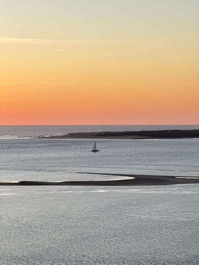 The image depicts a serene coastal scene at sunset, likely in La Teste-de-Buch, France, given the characteristic landscape of the Arcachon Bay region. The sky transitions from a pale yellow at the top to a vibrant orange near the horizon, with thin contrails from aircraft visible. A calm body of water, likely part of the bay, occupies the majority of the frame. In the middle distance, a small red and white sailboat navigates the water. Further in the background, a narrow strip of land with vegetation is visible, and on its shore, a white lighthouse stands out. In the foreground, a dark sandbar curves through the water, creating a distinct line across the scene. The overall impression is one of tranquility and natural beauty.