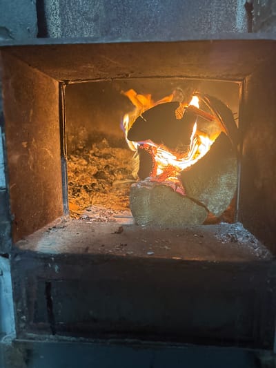 A close-up, eye-level shot shows a wood-burning stove in a rural setting, possibly in the Сароцкі сельскі Савет, Belarus. The focus is on the open firebox, where several logs are burning. Flames, in shades of orange and yellow, are visible, along with glowing embers and ash covering the bottom of the firebox. The interior of the stove appears to be constructed from metal and brick. The surrounding environment is not visible, but the scene suggests a cold weather setting due to the presence of a fire.
