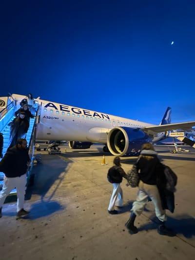 An Aegean A320neo aircraft is parked on the tarmac under a twilight sky. Several passengers are disembarking from a mobile stairway. The engine cowling of the plane is prominently displayed, along with the Aegean logo and "A320neo" printed on the fuselage. A crescent moon is visible in the upper left corner of the image. Some ground support equipment can be seen next to the plane. The photo appears to have been taken from an angle, possibly by someone on the ground.