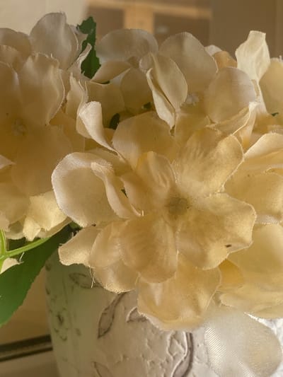 The image displays a close-up view of a bouquet of light yellow artificial flowers, possibly hydrangeas, with some green leaves visible. The flowers are made of fabric and have frayed edges, suggesting they are decorative. The bouquet is placed in a white vase or pot that appears to be textured and possibly hand-painted with subtle floral designs. The background is blurred, showing a warm, neutral-toned environment, possibly an indoor setting. There is no indication of the city of Важное, Russia, or any specific urban environment. The focus is entirely on the floral arrangement.