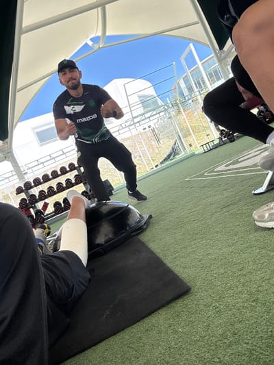A man in a Mazda shirt and cap stands with clenched fists in a gym. Dumbbells are lined up near him. A person sits in the foreground with a wrapped leg and a blue and white shoe. Green turf covers the floor, and a black mat is visible near the seated person. Outside, a white building is visible under a blue sky. The scene is partly shaded by a white awning.