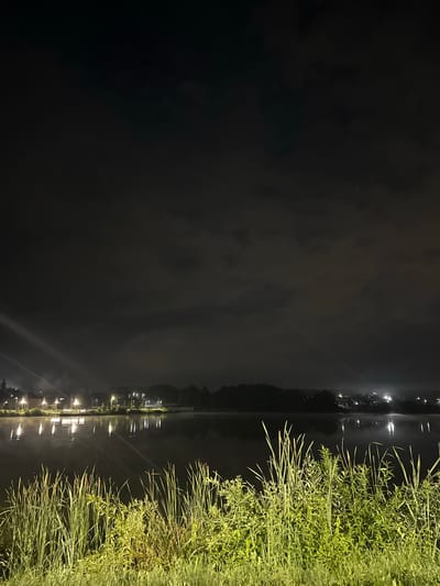 This is a nighttime photograph of a lake in the village of Zvanka, Russia. The foreground is dominated by tall, green grass and reeds. The lake's surface is dark and reflects the lights from buildings and street lamps visible on the opposite shore. These lights cast elongated, shimmering reflections on the water. The opposite shore features a line of trees and structures, some with illuminated windows. The sky is dark with scattered clouds, and a faint mist or fog appears to be settling over the lake. Two faint beams of light, possibly from spotlights or car headlights, are visible in the upper left and right portions of the sky, cutting through the darkness. There are no people or animals clearly visible in the scene. The overall atmosphere is serene and quiet.